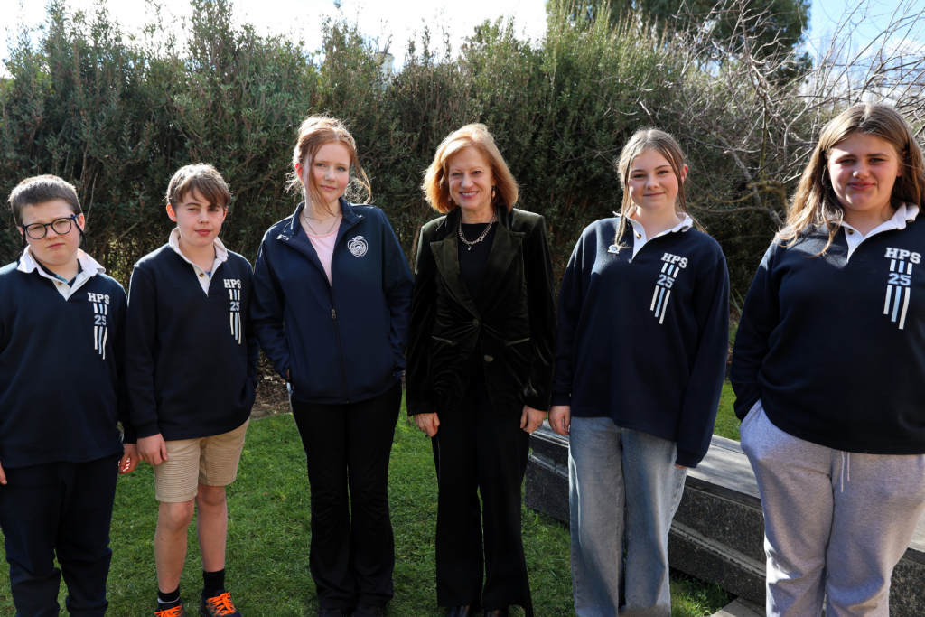 A group of five students in school uniforms stand outdoors next to an adult woman in a black outfit, smiling at the camera with greenery and a stone bench in the background.