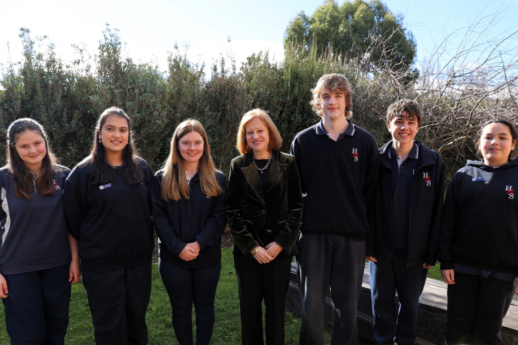 A group of six students in school uniforms stand outdoors beside an adult woman in a green jacket, all smiling at the camera with greenery and trees in the background.