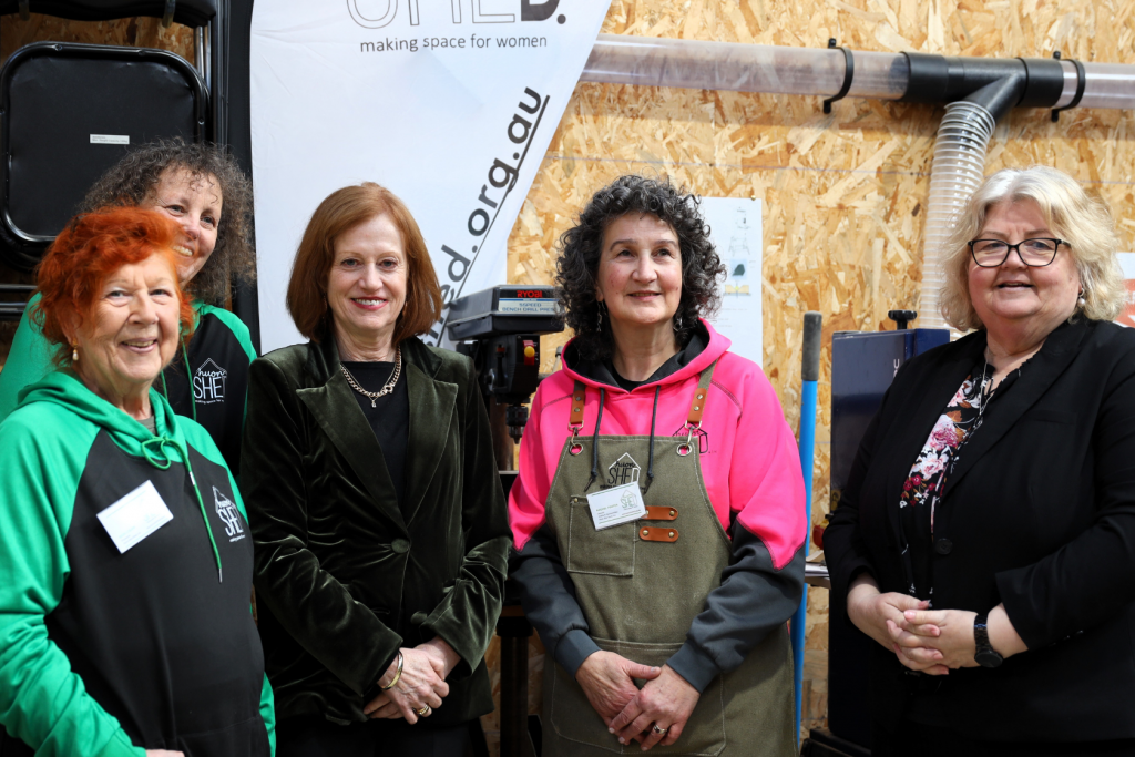 Five women stand together smiling indoors. One wears a black blazer, others wear hoodies or aprons. A banner behind them reads Shed: making space for women with a wood-panel wall and workshop equipment visible.