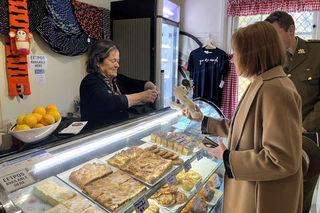 A woman behind a bakery counter hands a customer change. The customer, holding a small stuffed animal, stands next to a man in uniform. Pastries and baked goods are displayed in the glass case.