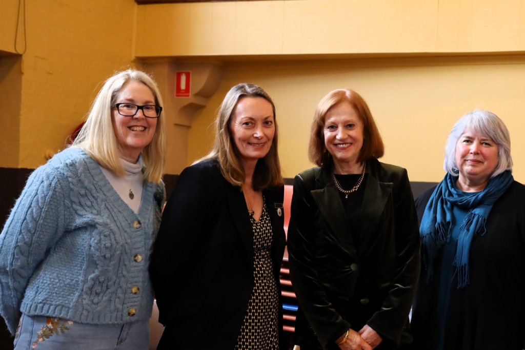 Four women standing side by side indoors, smiling at the camera. They are dressed in business casual and formal attire, against a yellow and brown background.