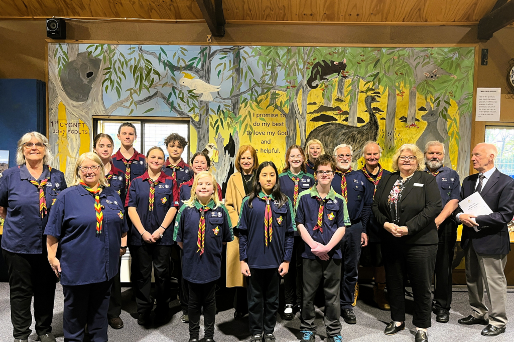 A group of scouts and adults pose indoors in front of a colorful mural with animals and text. Some wear scout uniforms with scarves; others are in formal attire. The mood is cheerful and formal.
