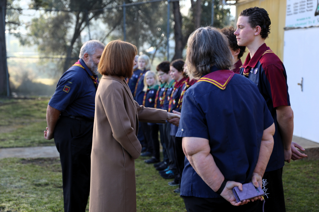 A woman in a tan coat shakes hands with a person in a scout uniform, while other scouts stand in a line outdoors on a grassy area. Trees and a building are visible in the background.