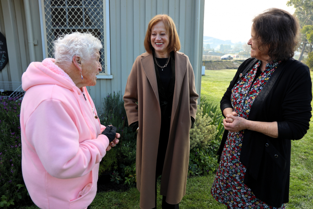 Three women stand outside talking and smiling; one wears a pink hoodie, another a brown coat, and the third a floral dress with a black cardigan. They are in front of a gray building on a grassy lawn.