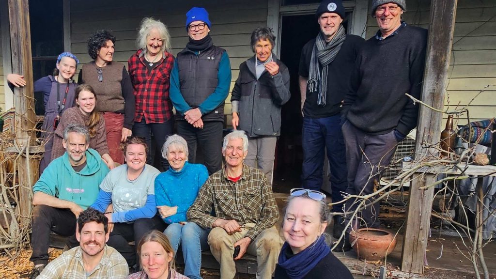 A group of fourteen adults, men and women, are smiling and posing together on the porch of a rustic wooden house. Some are sitting on the steps, while others stand behind them. The atmosphere is relaxed and cheerful.