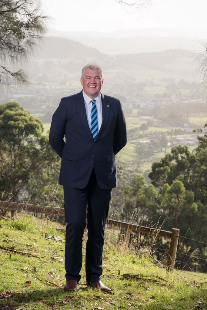 A man in a dark suit and striped tie stands smiling outdoors on grass with rolling hills, trees, and a town visible in the background under a hazy sky.