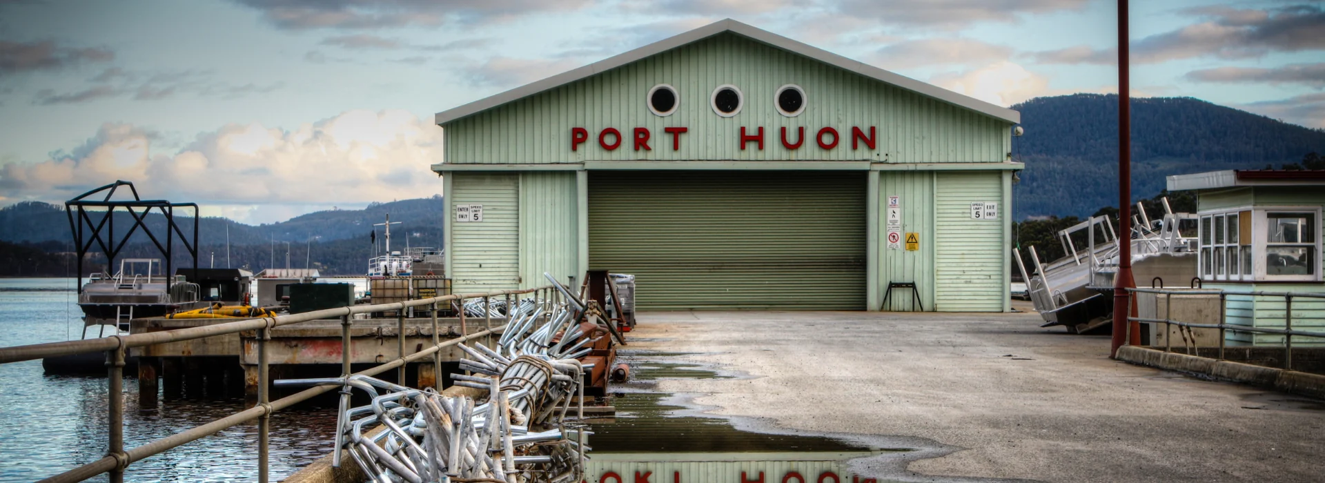 A pale green warehouse with PORT HUON in red letters on the front stands at the end of a pier, surrounded by water and boats, with mountains and cloudy skies in the background.