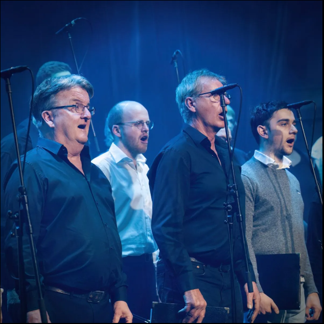 A group of men, standing and singing on stage in front of microphones, dressed in formal and semi-formal attire, illuminated by blue stage lighting.
