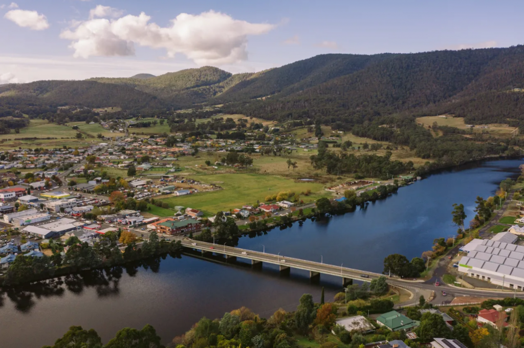 Aerial view of a small town by a river, with a bridge crossing the water, green fields, houses, and forested hills in the background under a partly cloudy sky.