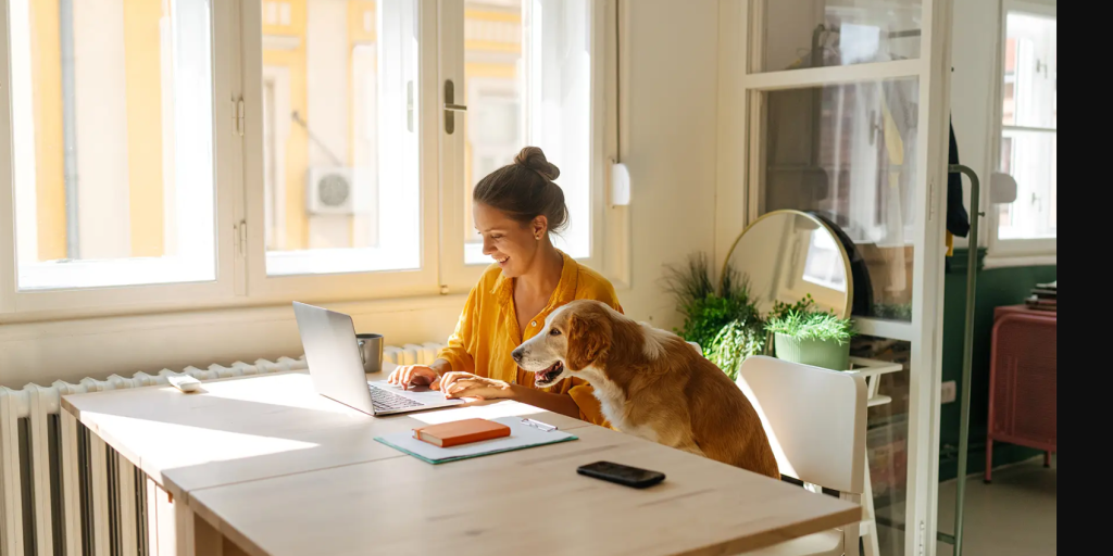 A woman in a yellow shirt smiles while working on a laptop at a desk in a bright room, with a dog sitting beside her looking at the screen.