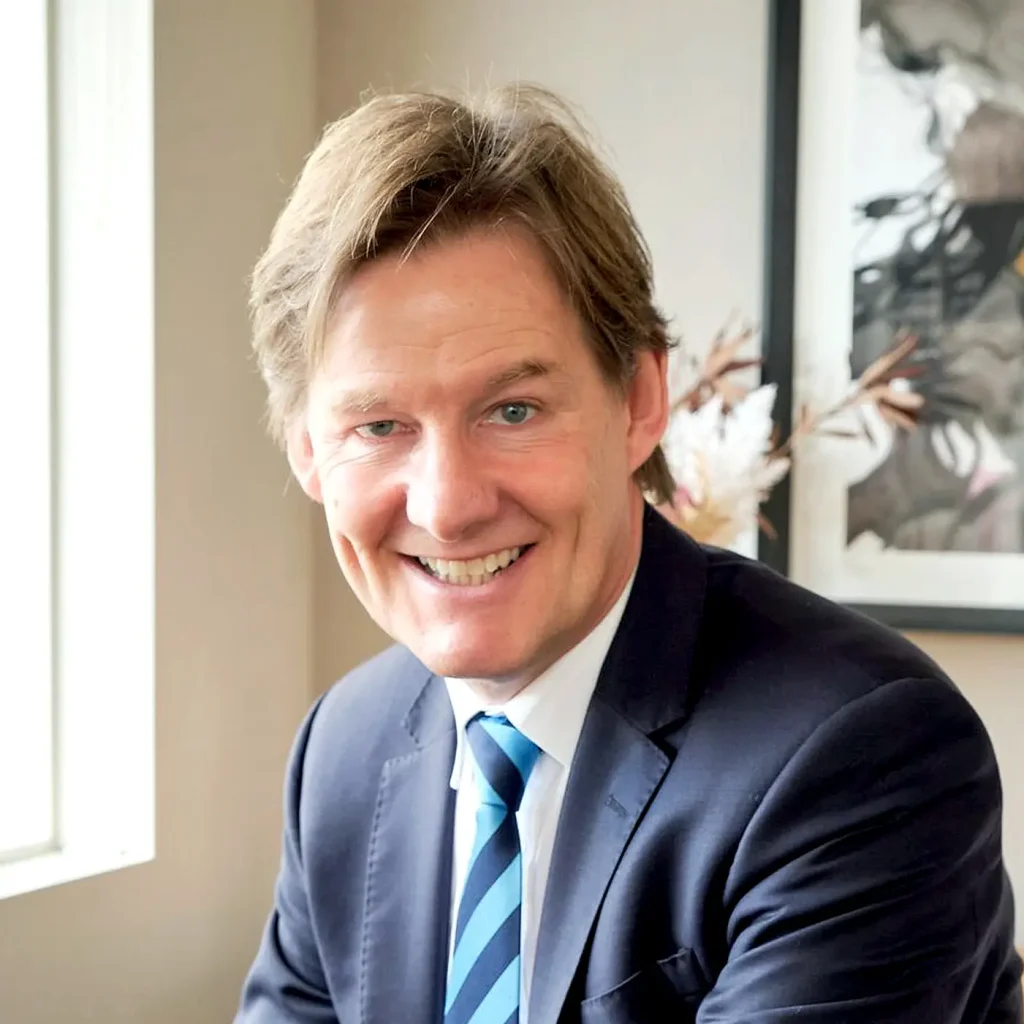 A man in a dark suit and blue striped tie smiles at the camera, sitting indoors near a window with natural light, with a framed picture and decorative plant in the background.