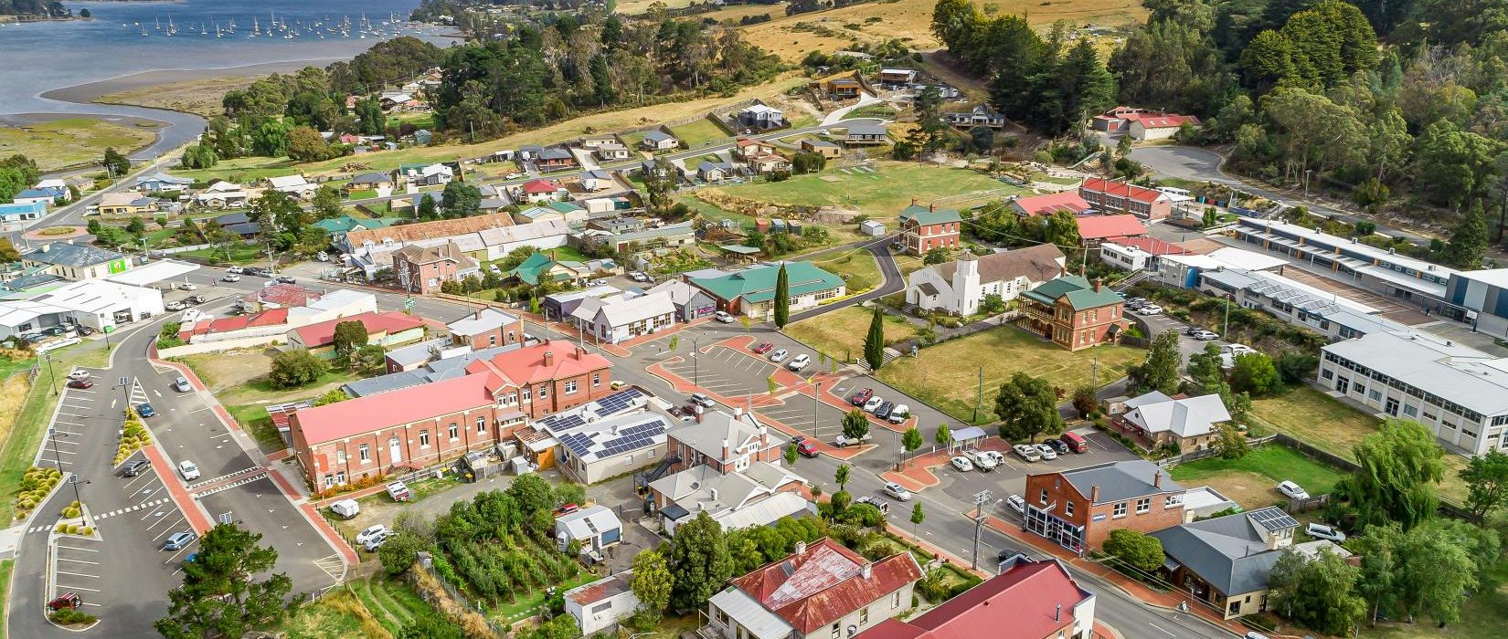 Aerial view of a small town with red-roofed buildings, green trees, winding roads, and a river with boats in the background, surrounded by rolling hills and open fields under a blue sky.