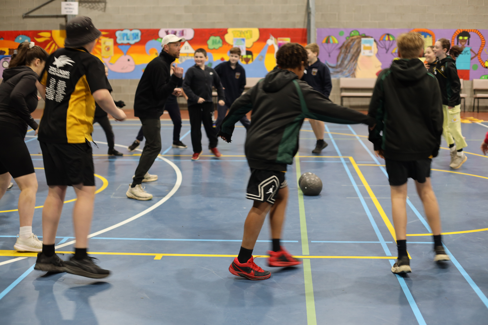 A group of students play a lively indoor soccer game in a gym with a colorful mural on the wall behind them. Everyone is in motion, focusing on a black soccer ball in the center of the action.