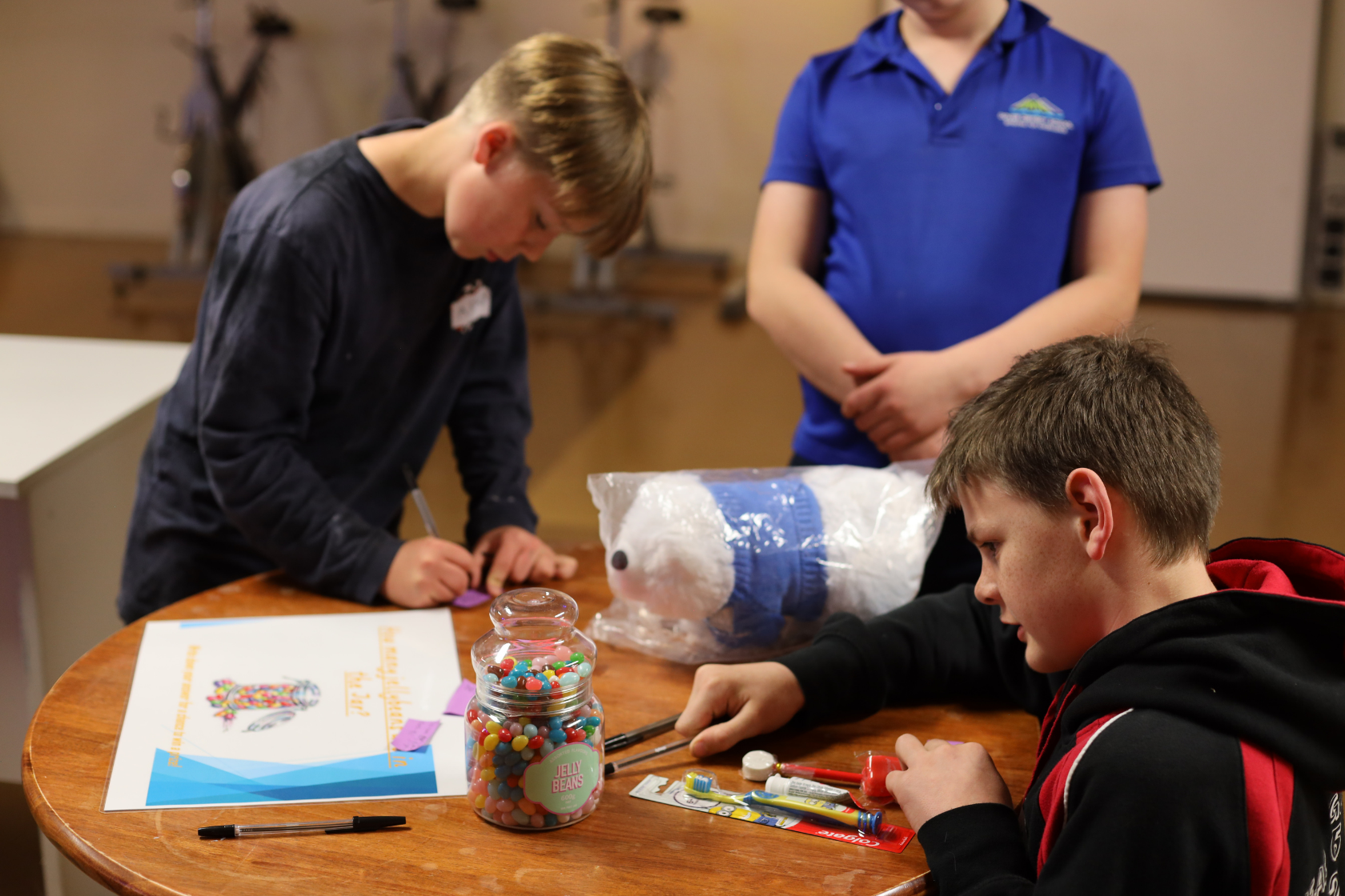 Three boys work at a round wooden table with craft supplies, a jar of colorful candy, and a plush toy. Two are focused on crafting, while the third stands nearby in a blue shirt. Exercise bikes are blurred in the background.