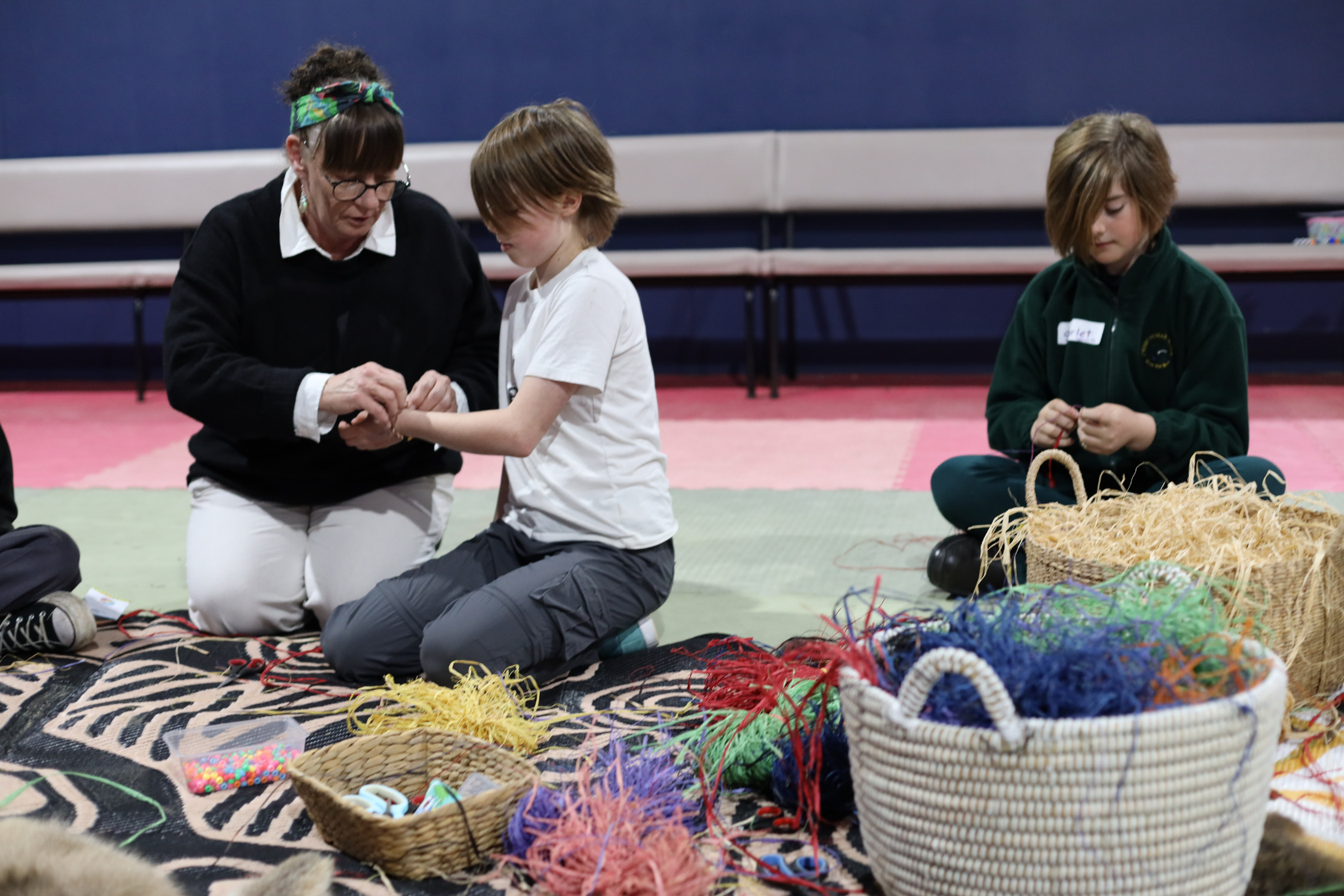 A woman helps a child with a craft project, while another child sits nearby weaving with colorful materials. Baskets filled with craft supplies are on the floor around them. They are seated on patterned mats indoors.