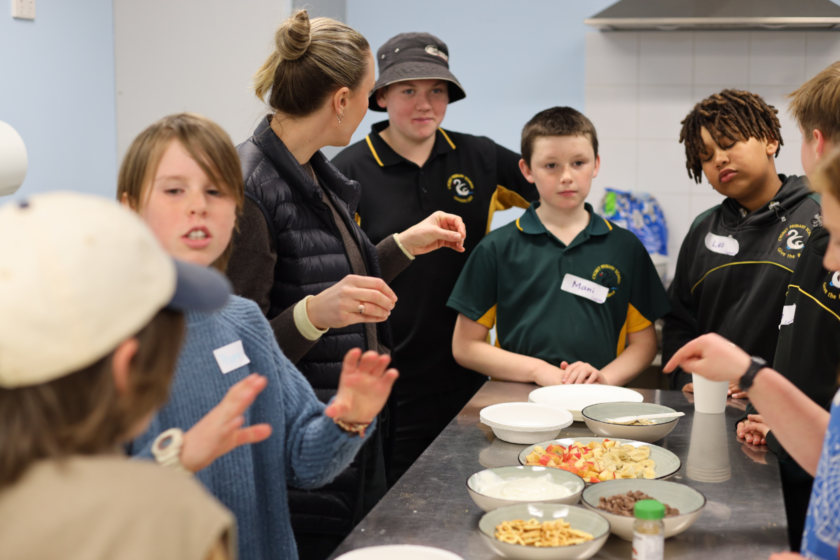 A group of children and an adult stand around a kitchen counter with plates of fruit and snacks, engaged in conversation and food preparation.