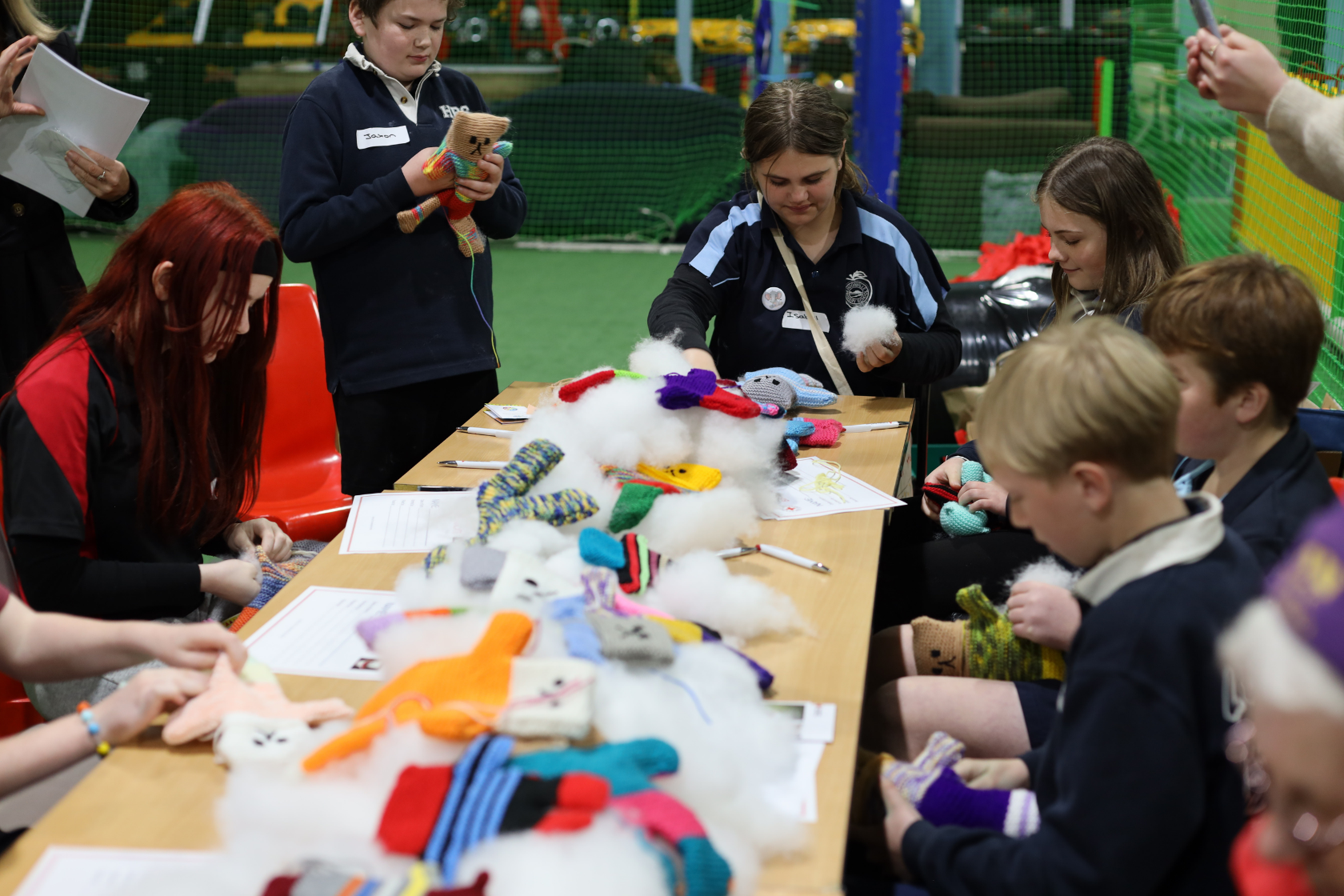 A group of children sit and stand around a table filled with colorful stuffed toys and white stuffing, working together on a craft project in an indoor recreational area.