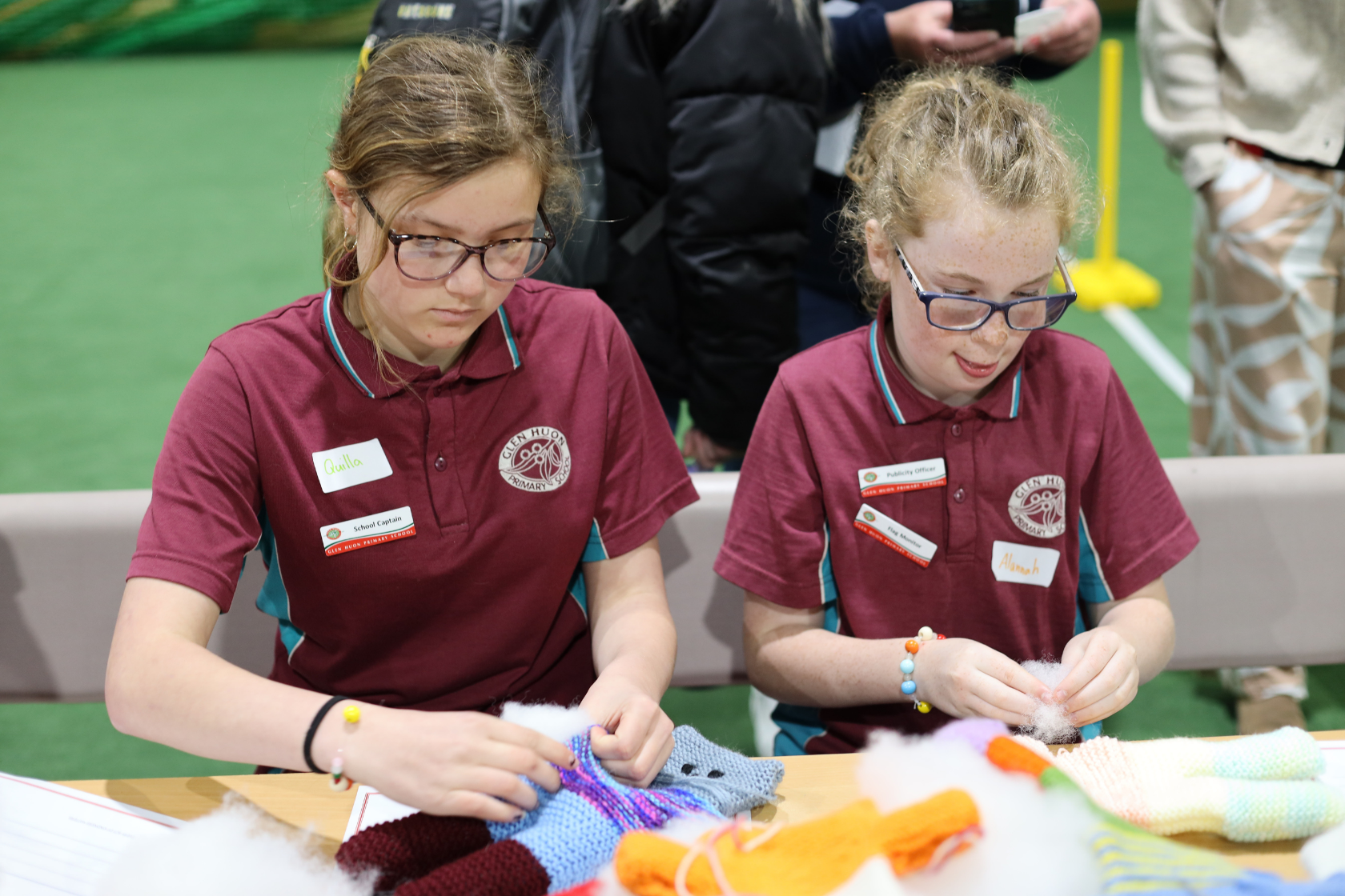 Two girls in maroon uniforms with name tags are sitting at a table, working with colorful yarn and stuffing. They appear focused on their craft project in an indoor setting with a green floor.