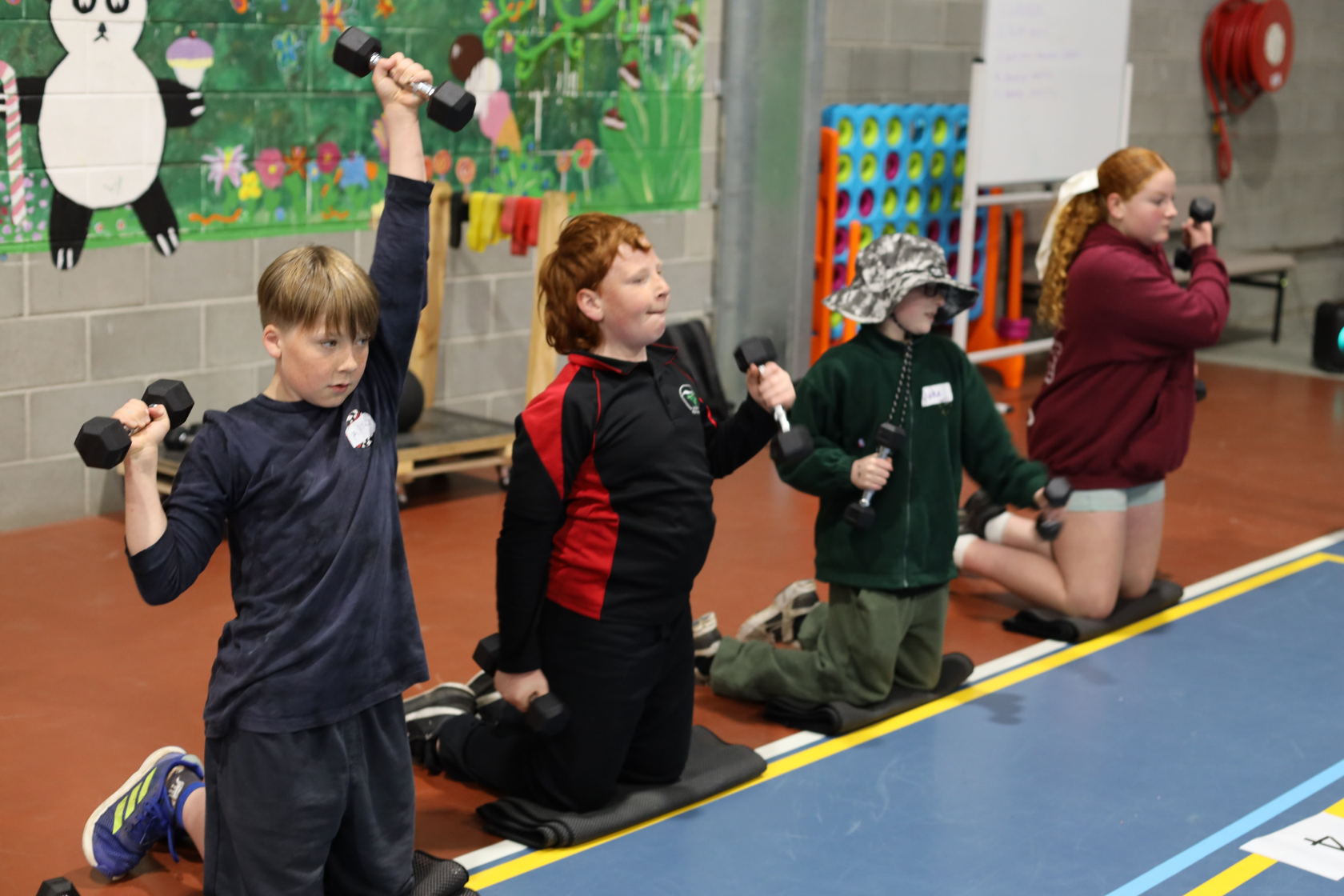 Four children kneel on mats in a gym, each holding a dumbbell in each hand and performing shoulder presses. A colorful mural and gym equipment are visible in the background.