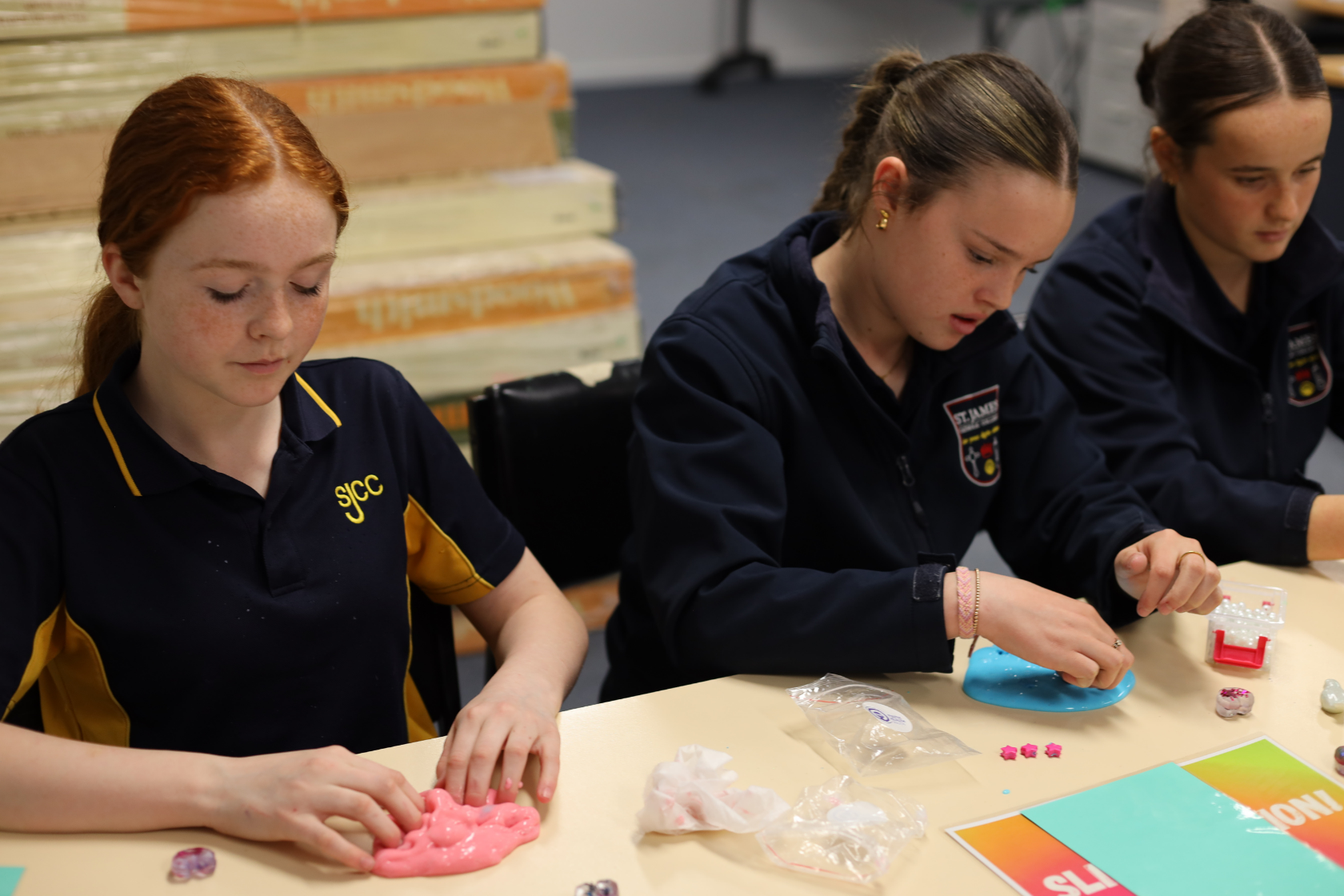 Three girls in school uniforms sit at a table making crafts with colorful clay and small decorative items. The table has craft supplies and instruction sheets on it.