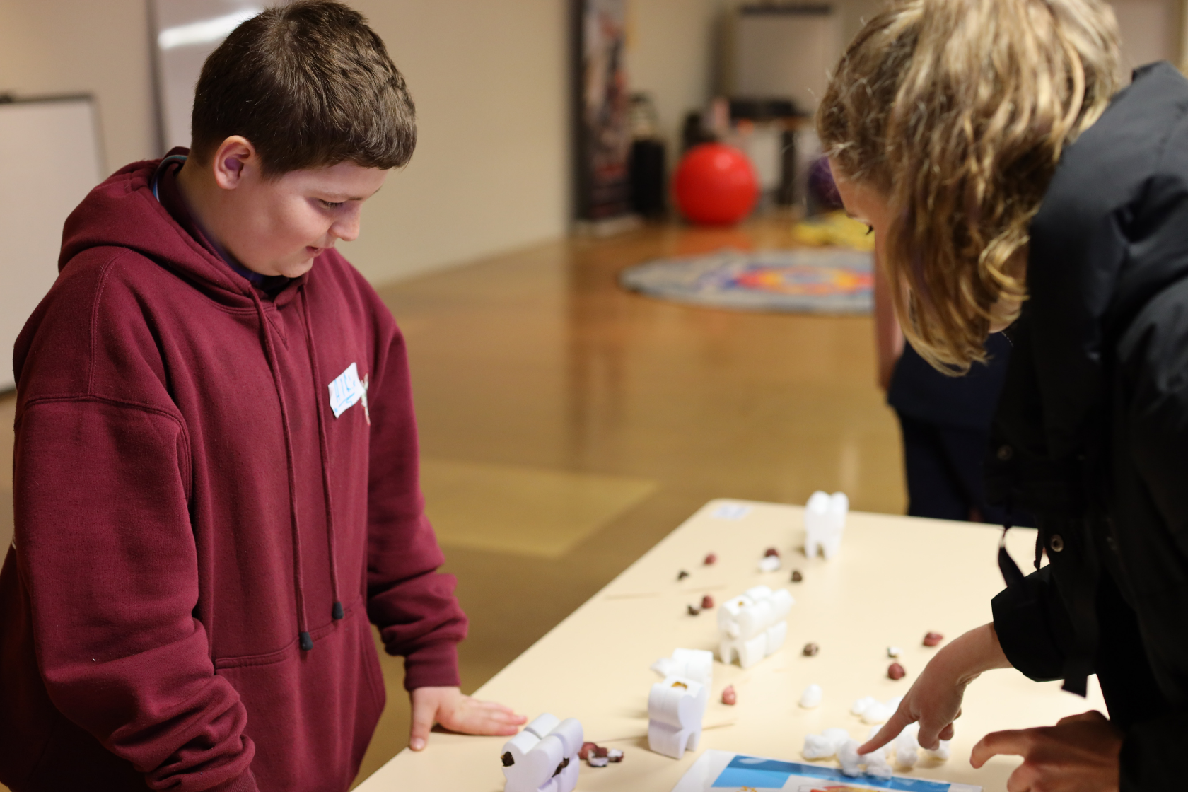 A boy in a maroon hoodie looks at a table with small white models, while an adult points at something on the table. The background is blurred, showing a spacious, indoor setting.