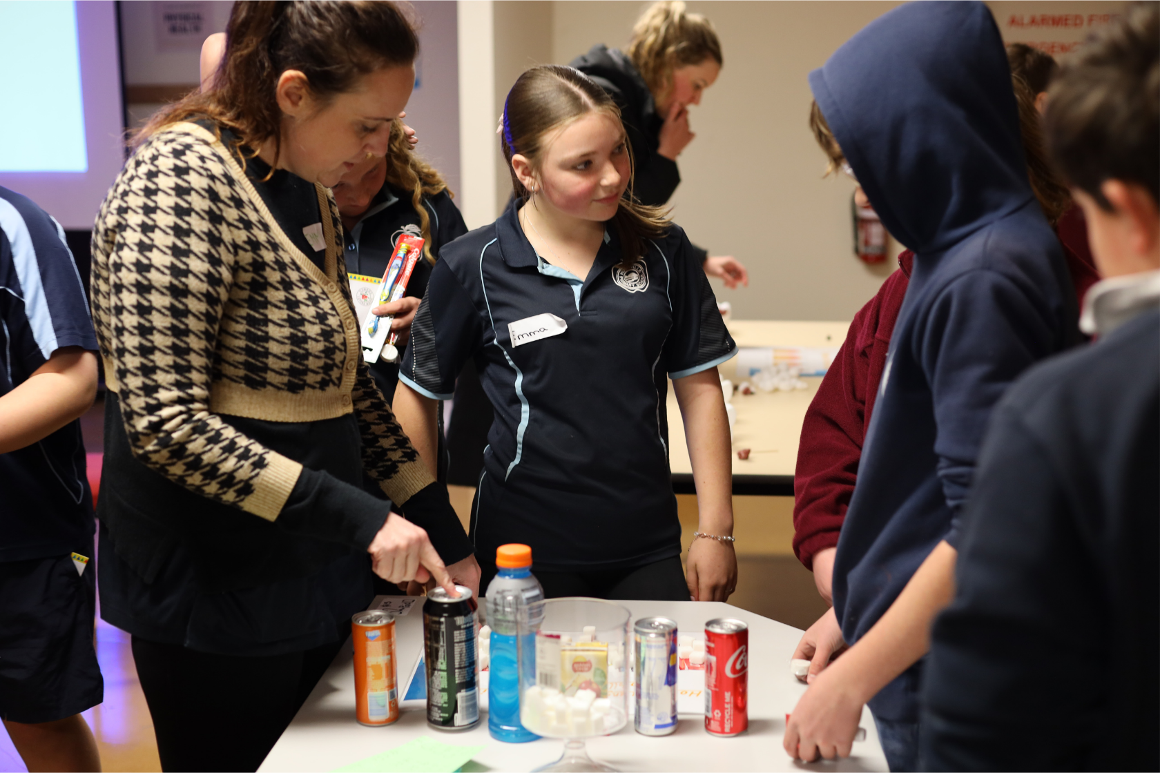 A group of students and an adult gather around a table with various drinks and sugar cubes. The adult points at the table, and the students look on attentively. The setting appears to be a classroom or educational event.