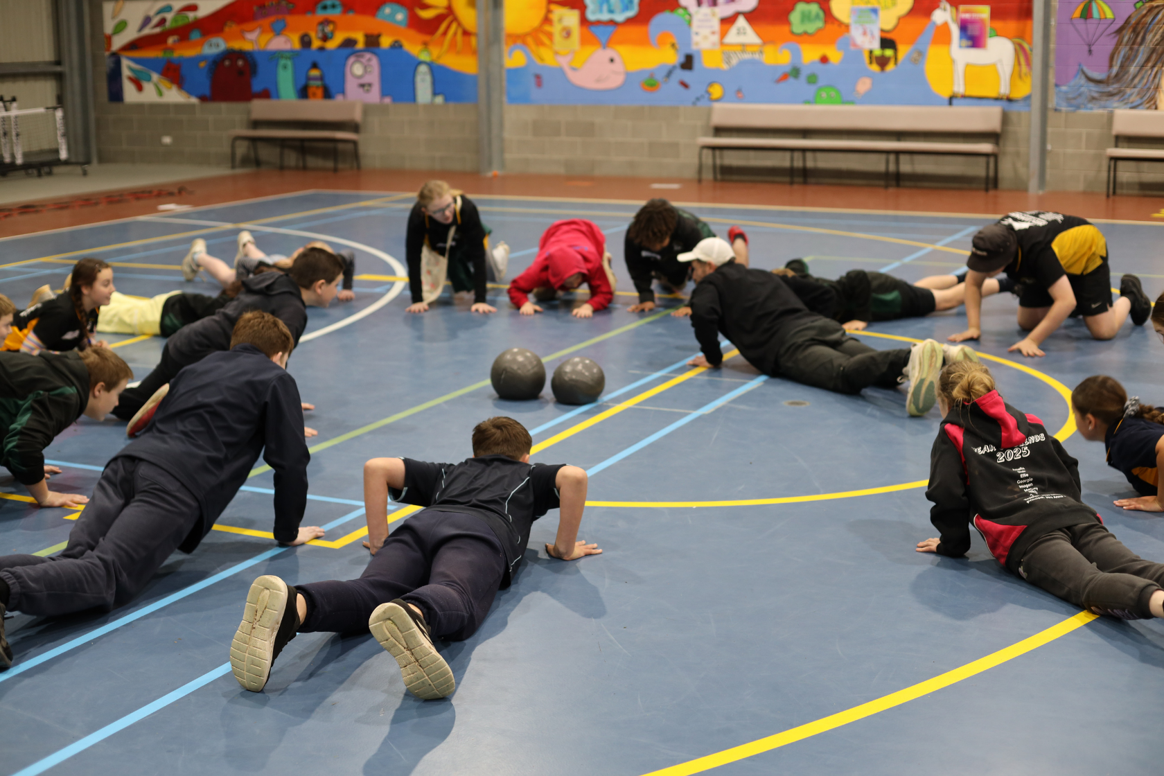 A group of children and adults are doing push-ups together on a gymnasium floor, with two medicine balls nearby. A colorful mural and benches are visible on the wall in the background.