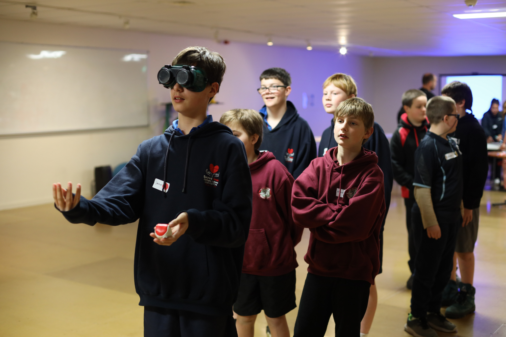 A group of boys stand in line indoors. The boy at the front wears goggles and reaches out with one hand, holding a small object in the other, while others watch and wait their turn.