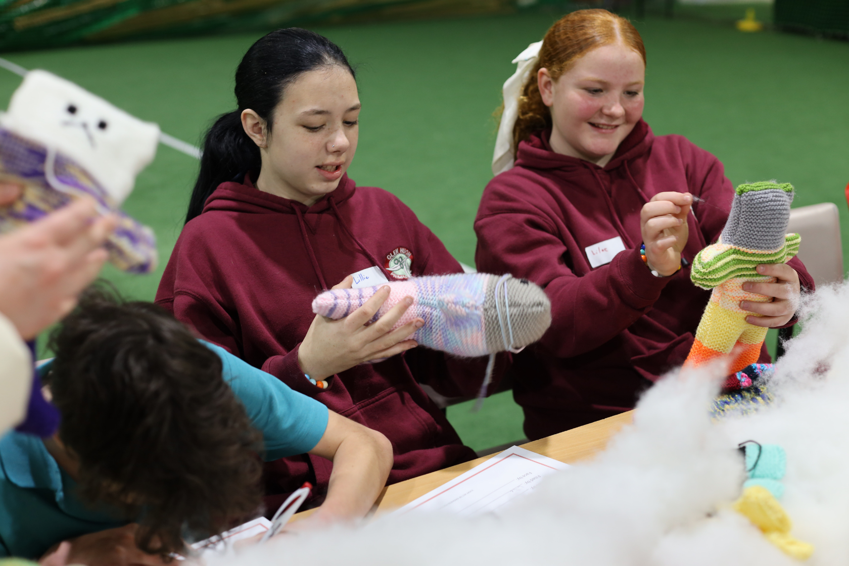 Two girls in maroon hoodies sit at a table, smiling and working on colorful knitted toys. They are surrounded by stuffing and other craft materials, participating in a group activity in a bright indoor setting.