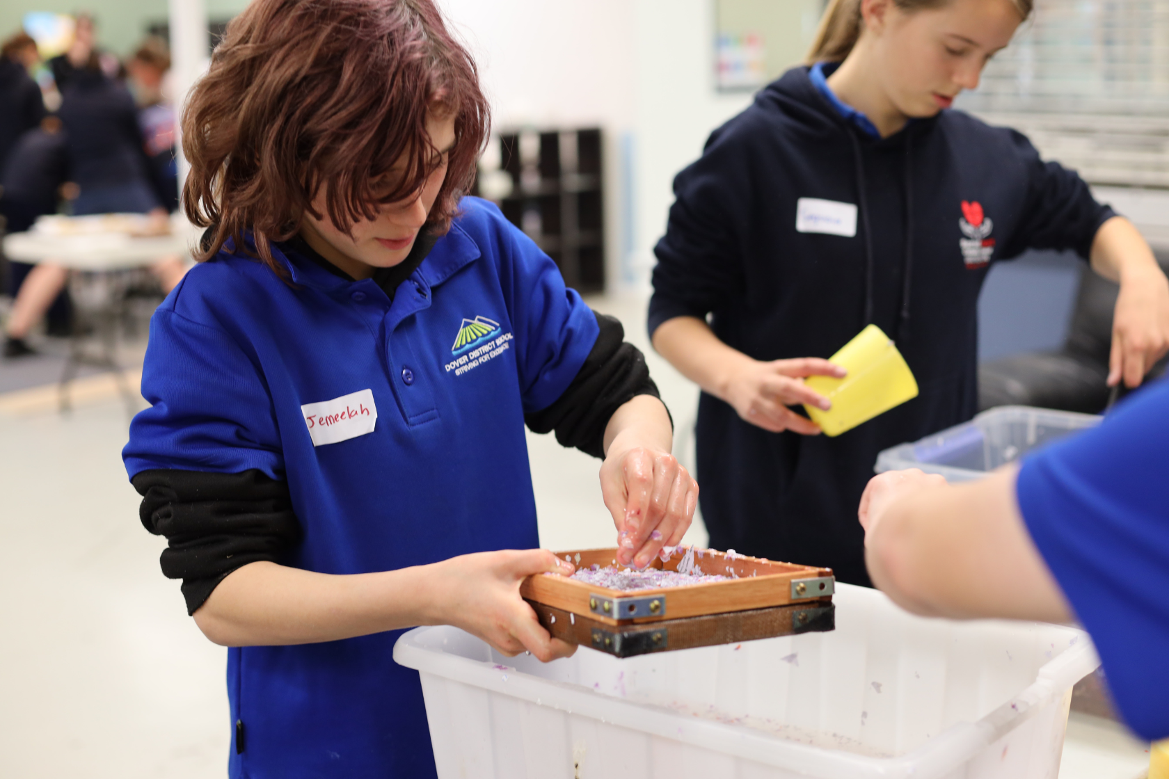 A child wearing a blue shirt and name tag uses a wooden frame to make paper at a white bin, while another child in a hoodie holds a yellow sponge nearby.