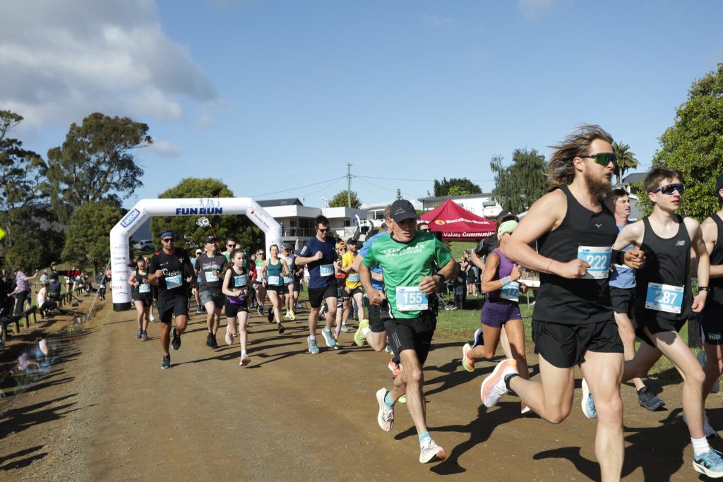A group of runners with race bibs begin a fun run on a dirt path under a white inflatable arch, with trees, tents, and spectators in the background on a sunny day.