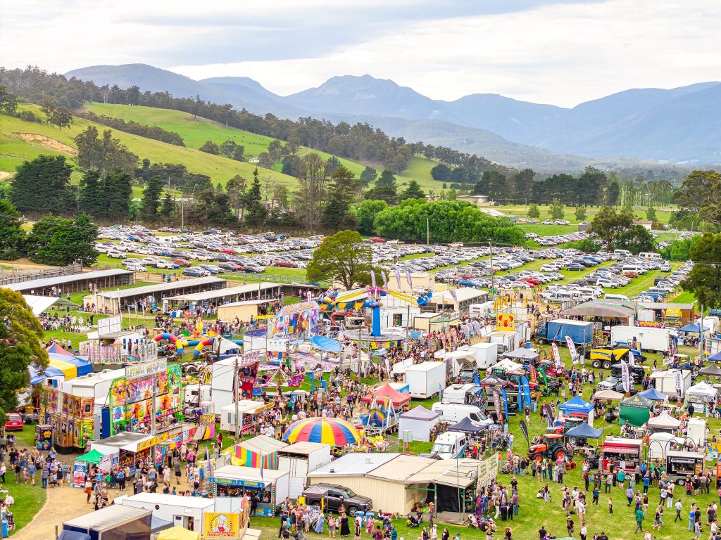 A large outdoor fair with colorful booths, rides, and food stalls is bustling with people. Cars fill the parking area in the background, with green hills and mountains under a cloudy sky beyond.