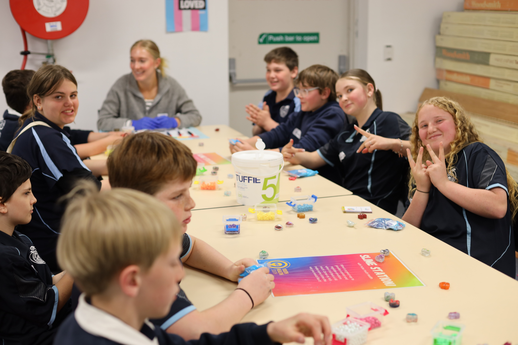 A group of children sit around a table in a classroom, engaging in a craft activity with colorful beads. An adult supervisor sits with them, and everyone appears to be enjoying the activity.