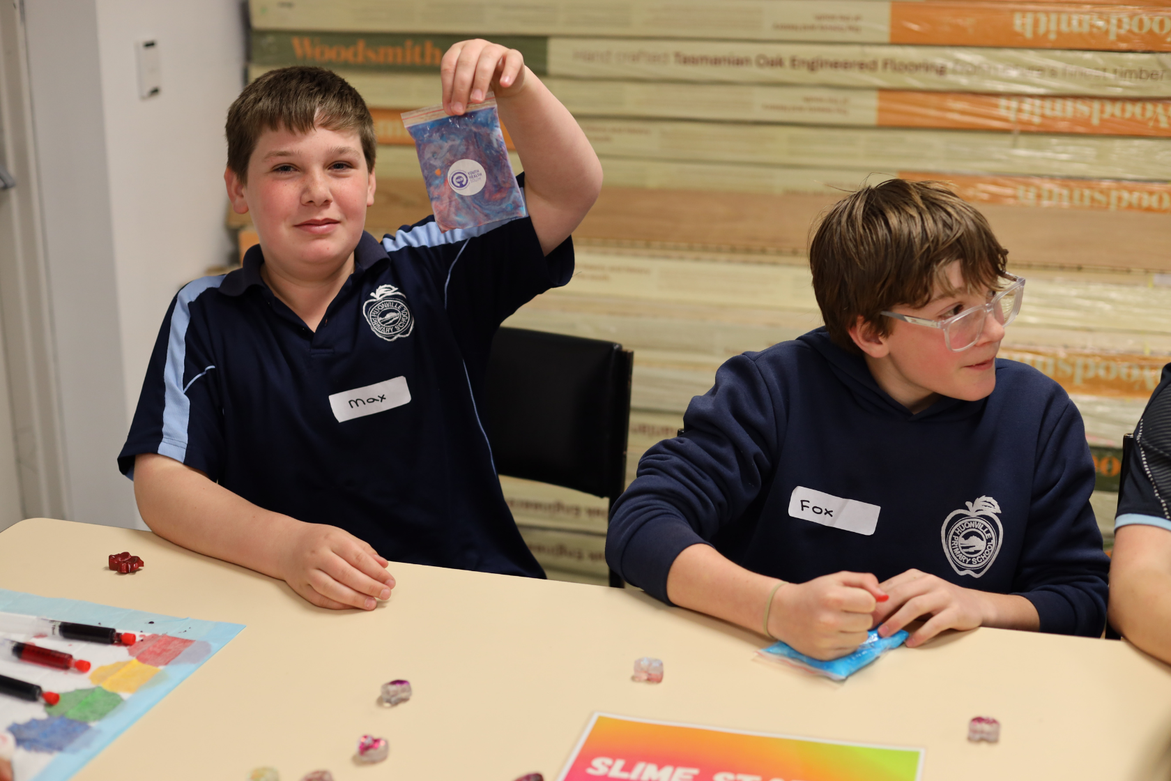 Two boys sit at a table making slime. One boy, wearing a dark blue shirt with Max on his name tag, holds up a small slime bag and smiles. The other boy, Fox, wearing glasses, looks down at his work.