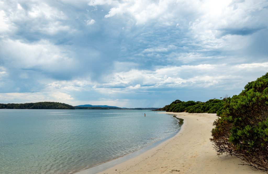 A sandy beach curves along calm, blue water under a cloudy sky, with green bushes lining the shoreline and distant hills visible across the bay.