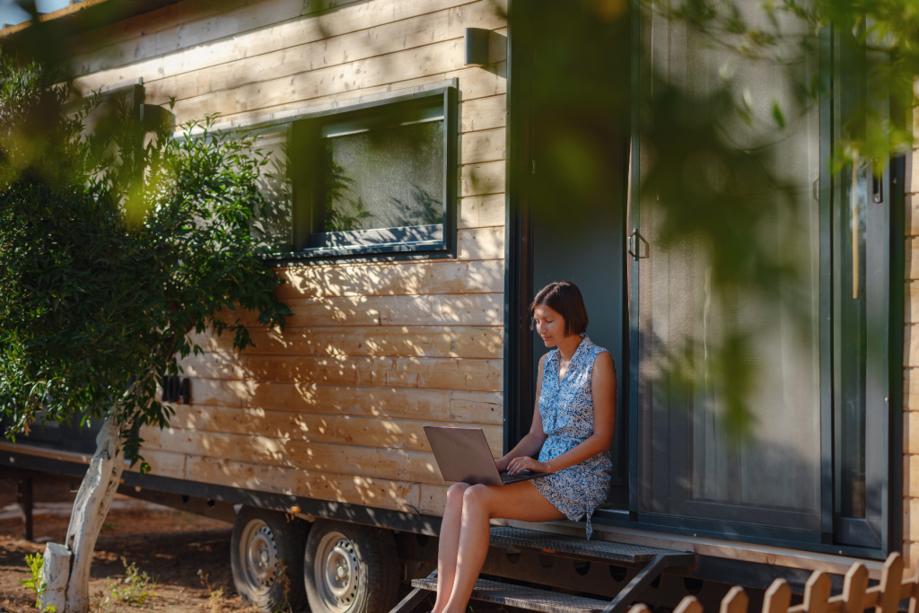 A woman in a blue dress sits on the steps of a wooden tiny house, working on a laptop. The house is surrounded by trees and sunlight, creating a peaceful outdoor atmosphere.