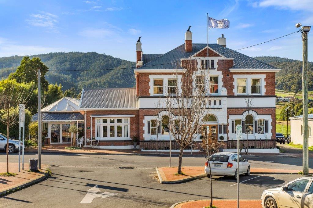 A large, two-story brick building with white trim and a flag on top stands at a street corner, surrounded by a few parked cars, with trees and hills in the background under a blue sky.