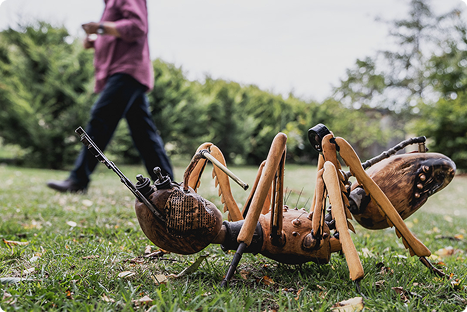 A large, wooden sculpture of an ant is on the grass in a park, with a person walking in the blurred background.