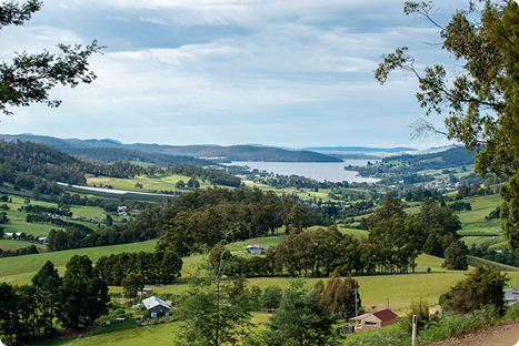 A scenic view of rolling green hills, scattered houses, and clusters of trees, with a wide river or lake in the background under a partly cloudy sky.