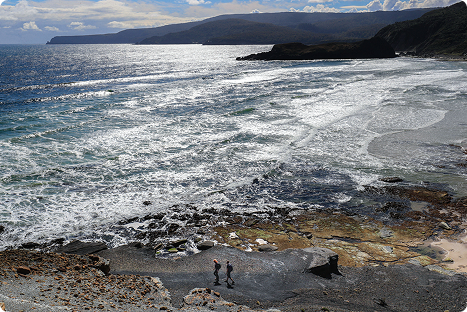 Two people walk along a rocky coastline with waves crashing onto the shore under a partly cloudy sky, with distant hills visible across the water.