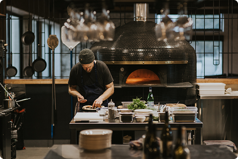 A chef prepares food at a kitchen counter in front of a large brick pizza oven, surrounded by cooking utensils, ingredients, and various kitchen tools.