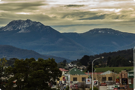 Town buildings and shops in the foreground with streetlights, framed by layered hills and tall mountains in the background under a cloudy sky.