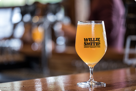 A stemmed glass filled with a hazy golden drink sits on a wooden bar. The glass is labeled Willie Smith’s Huon Valley, TAS. The background is softly blurred, showing a bar setting.