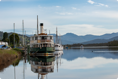 A vintage steamboat is docked on a calm river with its reflection visible in the water. Sailboats and mountains are in the background under a partly cloudy sky.
