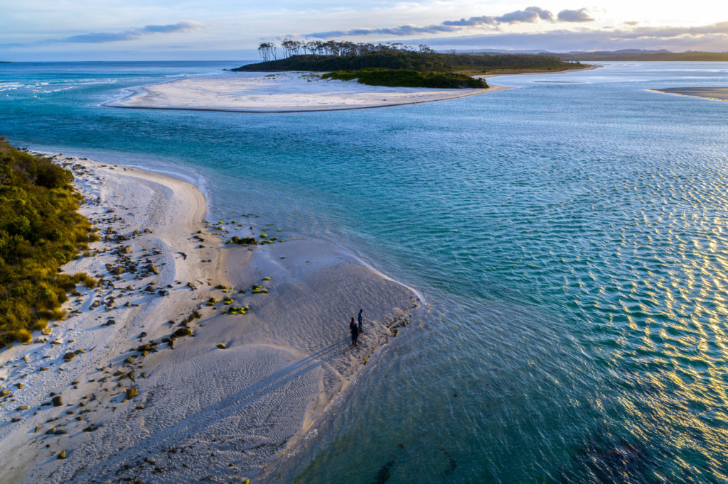 Aerial view of a person walking on a sandy beach surrounded by shallow blue water and small islands, with grass and rocks along the shoreline under a partly cloudy sky at sunset.