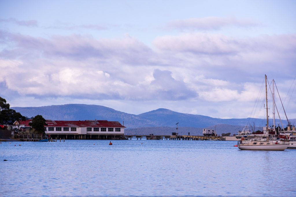A calm waterfront scene with a sailboat anchored on the right, buildings with red roofs on the shore, and mountains in the background under a partly cloudy sky.