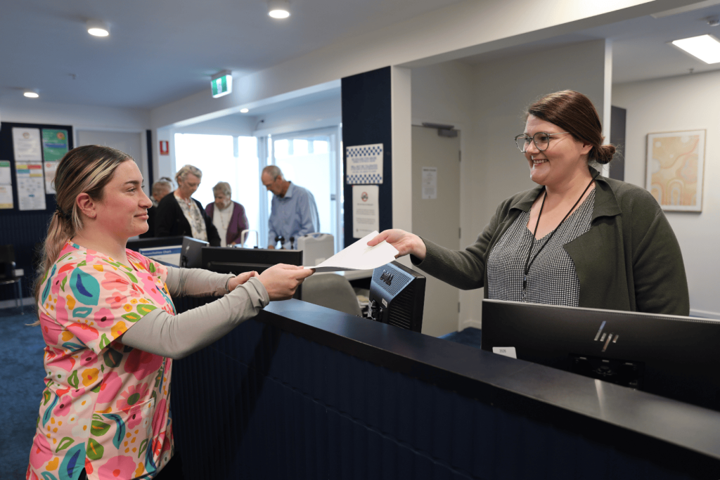 A woman in colorful scrubs hands paperwork to a smiling receptionist behind a counter, while several people wait in line in the background. The setting appears to be a medical or healthcare office.
