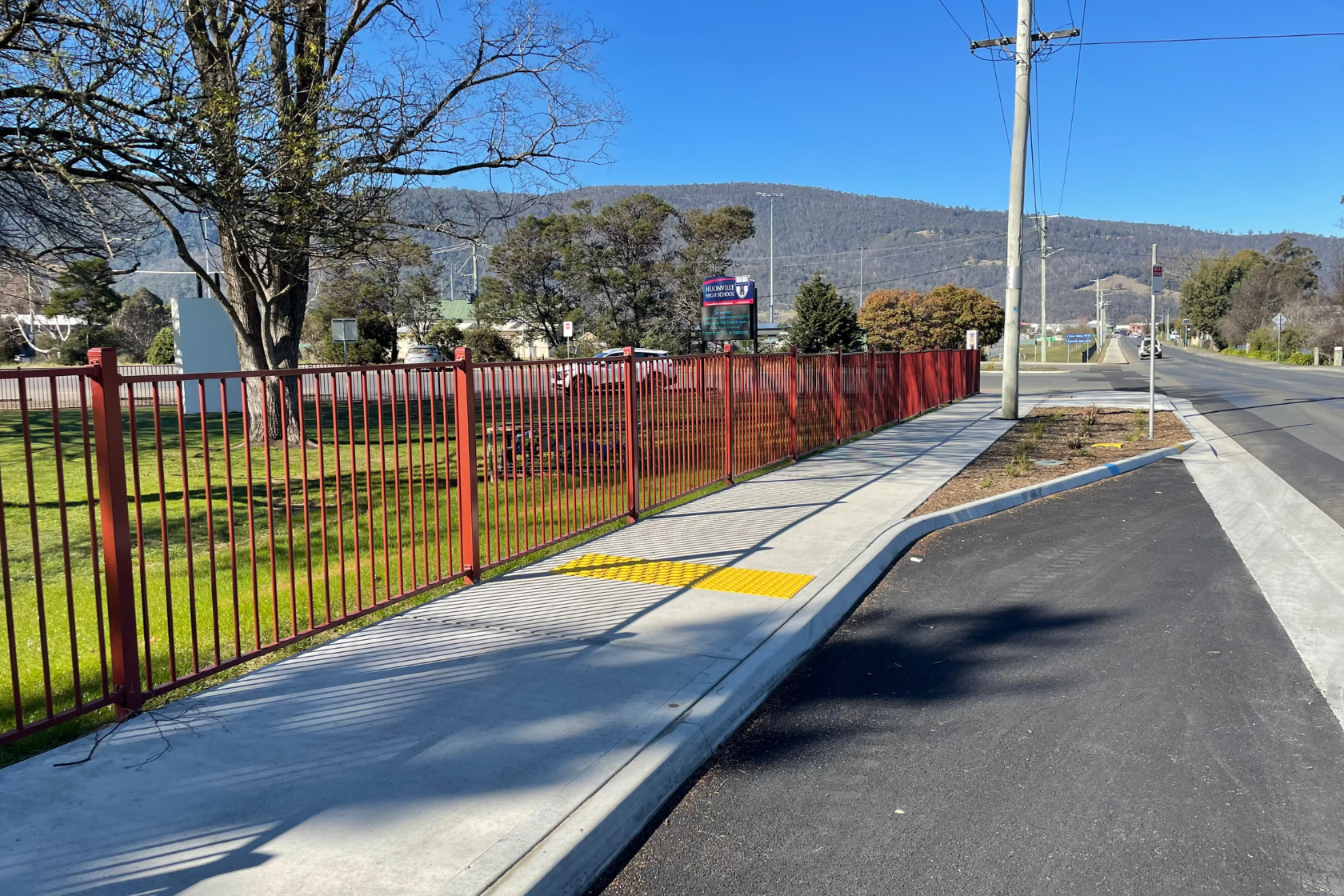 A sidewalk with a red fence runs beside a road on a sunny day. There is a yellow tactile paving section for accessibility, green grass, trees, and hills in the background under a clear blue sky.