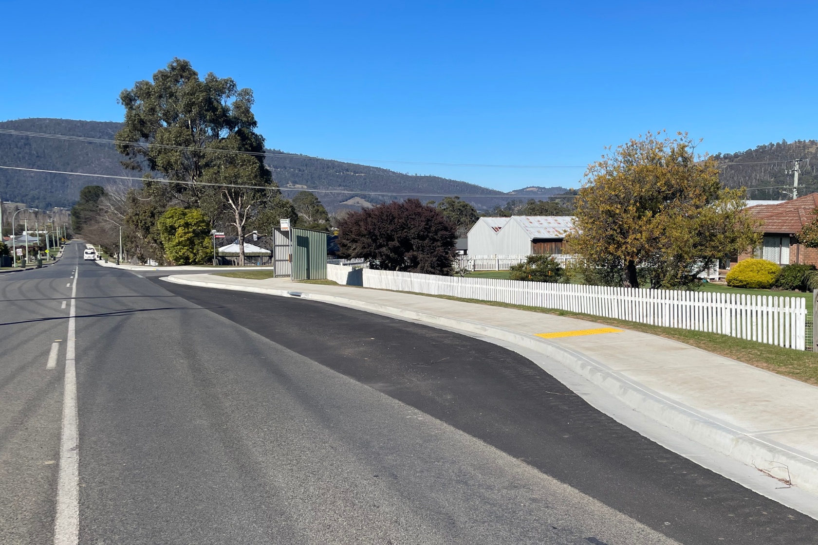 A wide, empty suburban road curves next to a sidewalk and white picket fences, with houses, trees, and mountains visible under a clear blue sky.