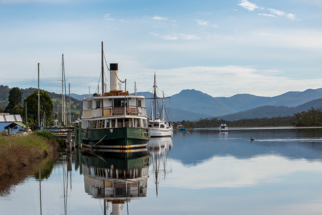 A vintage steamship is docked on a calm river, surrounded by sailboats. Trees, hills, and mountains are visible in the background under a partly cloudy sky, all reflected in the still water.